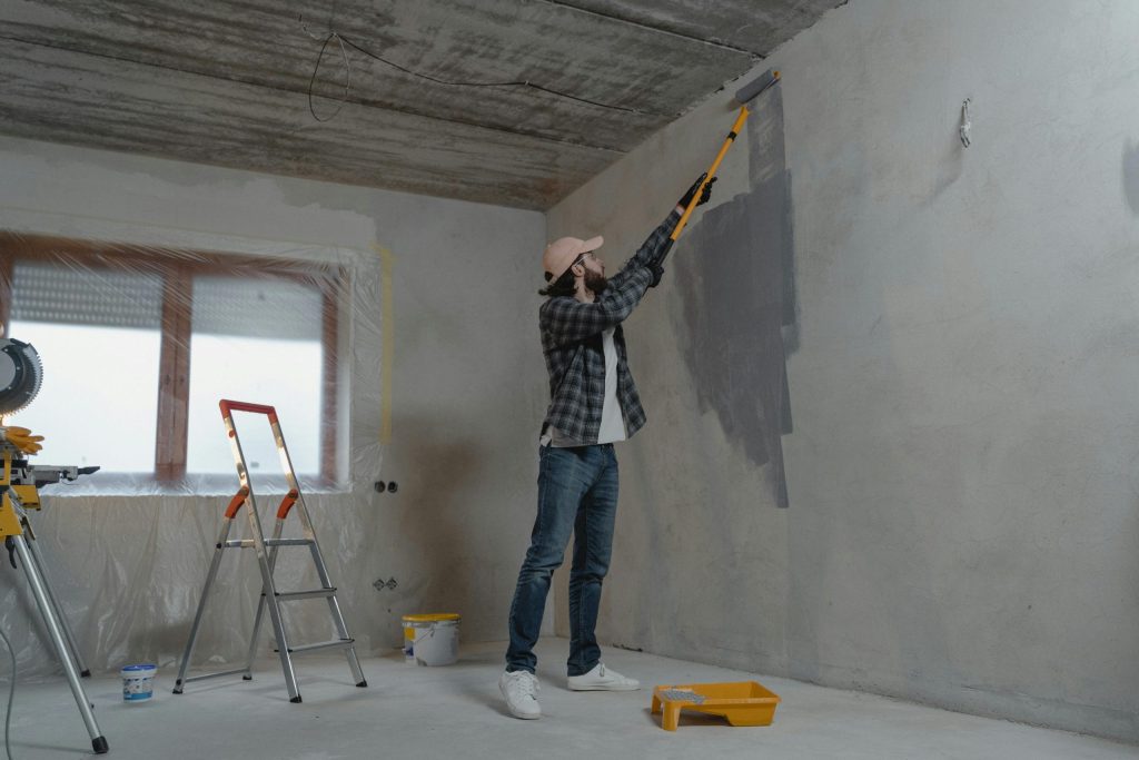 Man painting a wall with roller in a room; ladder and tools visible, home renovation scene